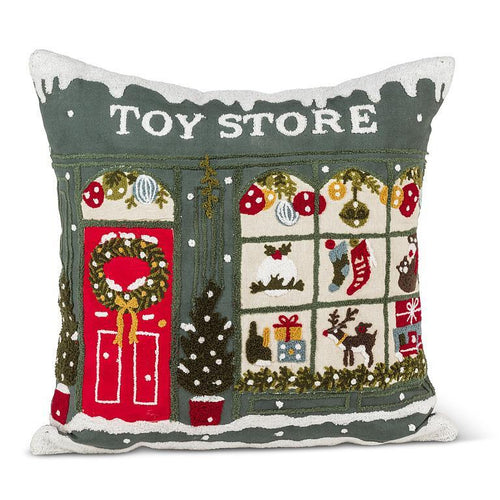 Square pillow with tufted toy-storefront scene—green facade, red door, wreaths, ornaments, and gifts in warm holiday tones.